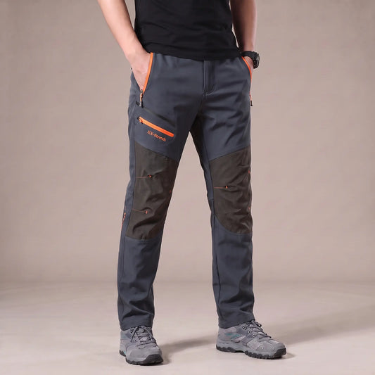 Men's grey stretch hiking trousers with dark grey knee panels and orange zip pockets, worn with hiking shoes