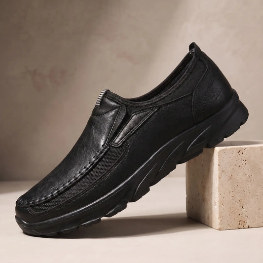 Stitched Apron Loafers in black, a slip on moccasin loafer with elastic side panels and a chunky black bottom, on a travertine display block