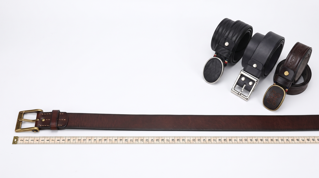 A men's leather belt uncoiled beside a measuring tape on a white studio surface