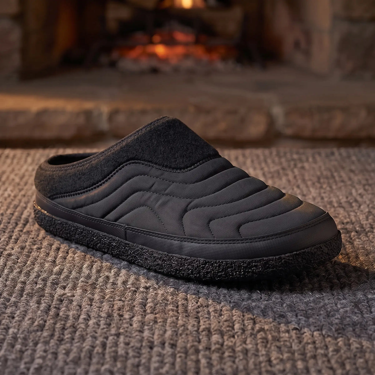 Men's black quilted backless slipper with felt collar and dark speckled sole on a wool rug by a fireplace