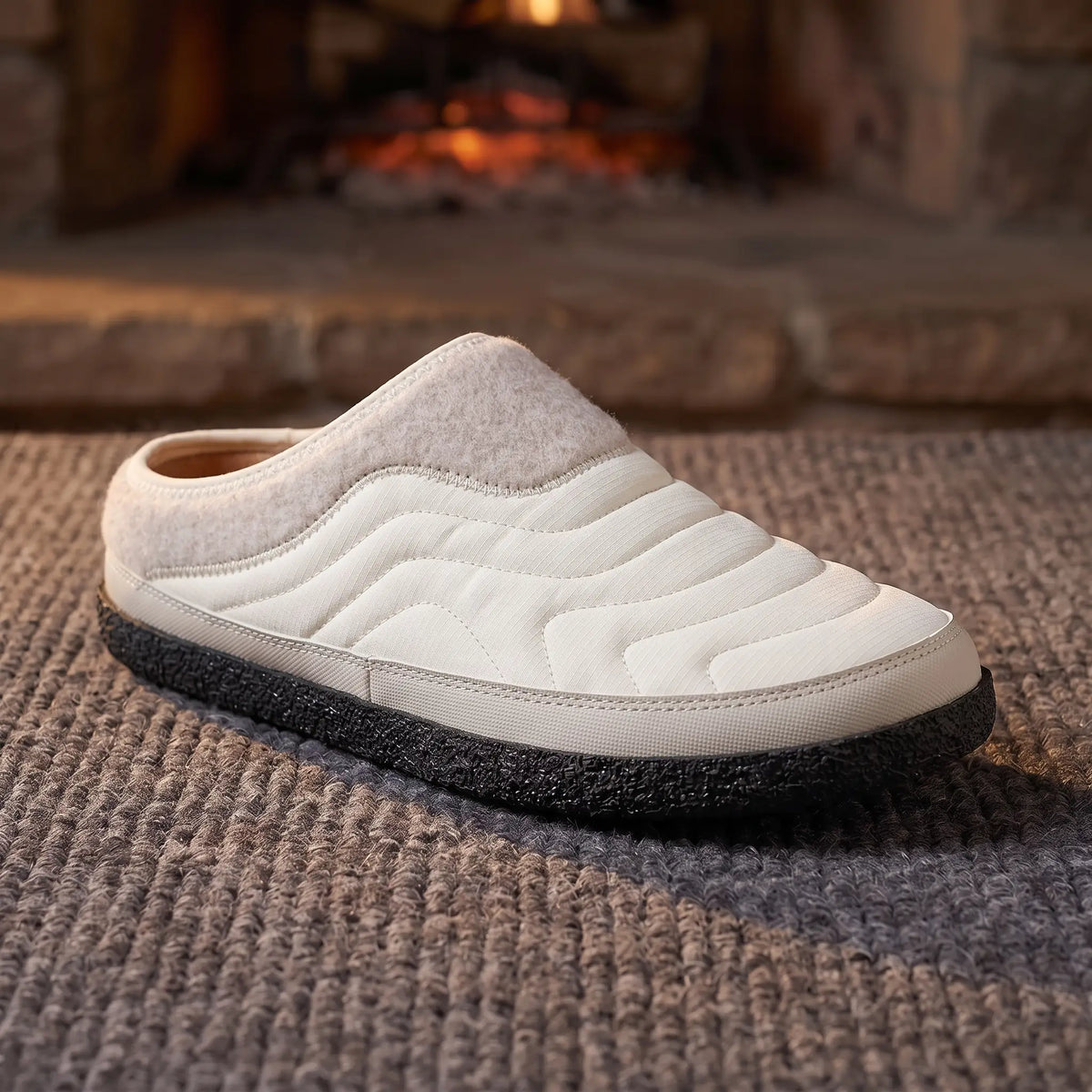 Men's white quilted backless slipper with felt collar and dark speckled sole on a wool rug by a fireplace