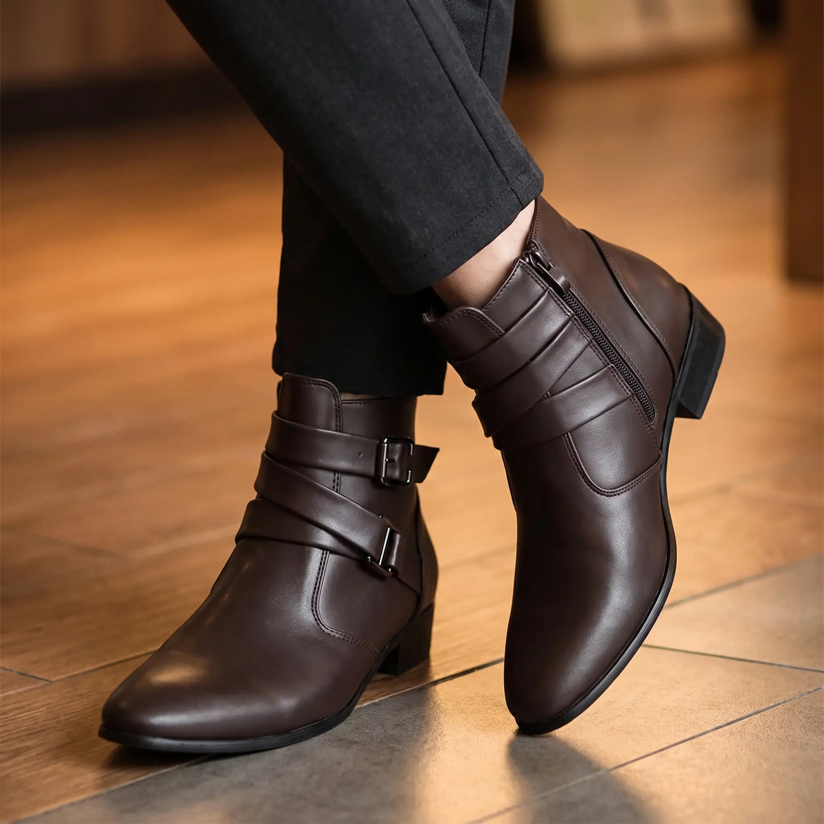Man seated with ankles crossed wearing brown pointed toe buckle ankle boots with dark trousers