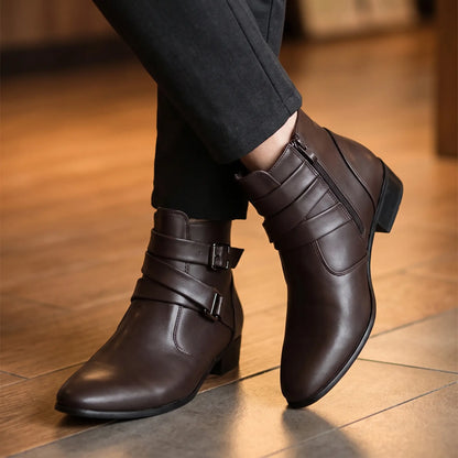 Man seated with ankles crossed wearing brown pointed toe buckle ankle boots with dark trousers