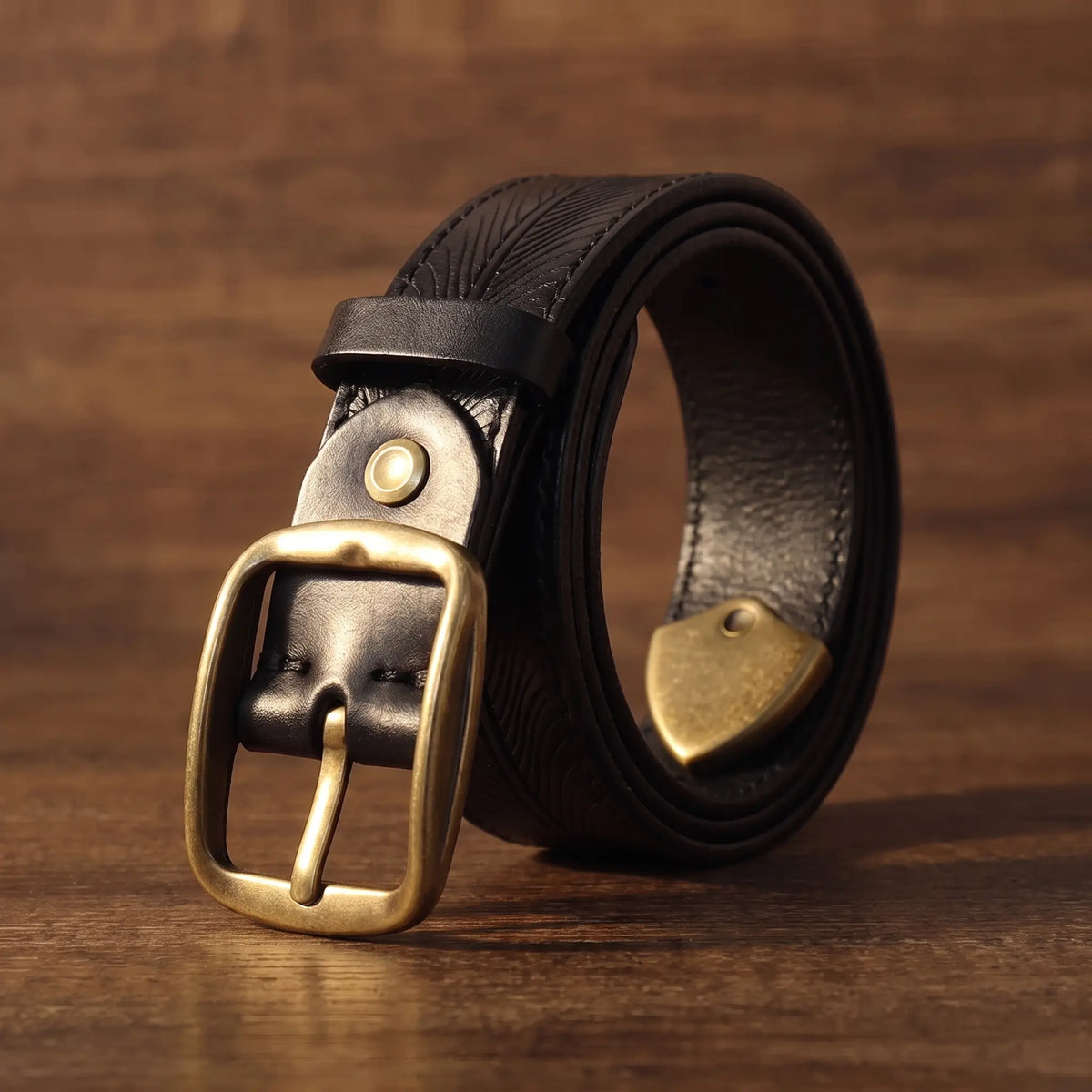 Men's black cowhide belt with feather embossed pattern and antique brass buckle, coiled on walnut