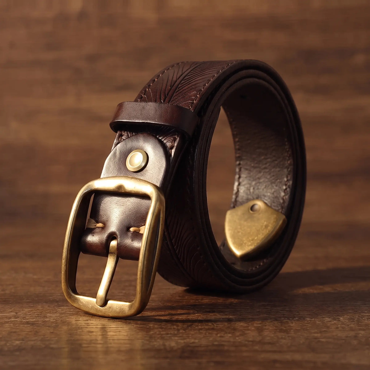 Men's brown cowhide belt with feather embossed pattern and antique brass buckle, coiled on walnut