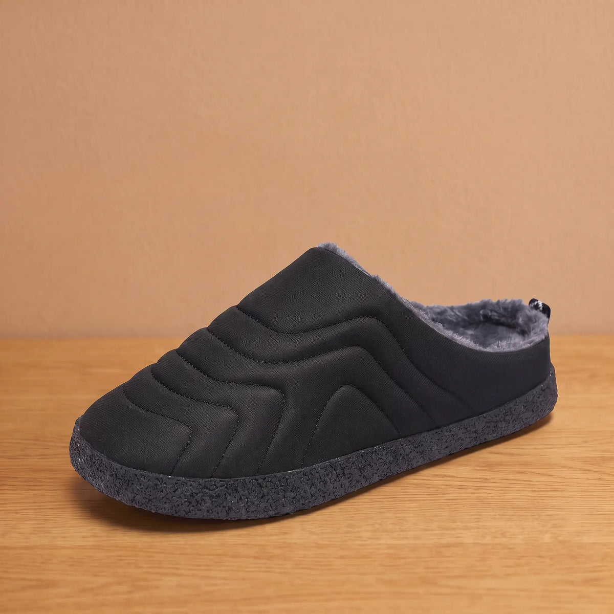 Men's black quilted backless slipper with plush faux-fur lining and speckled sole on an oak floor