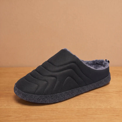 Men's black quilted backless slipper with plush faux-fur lining and speckled sole on an oak floor