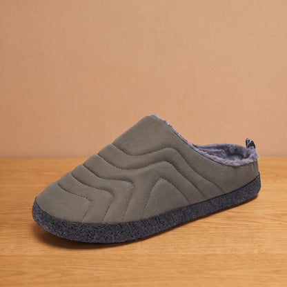 Men's grey quilted backless slipper with plush faux-fur lining and speckled sole on an oak floor