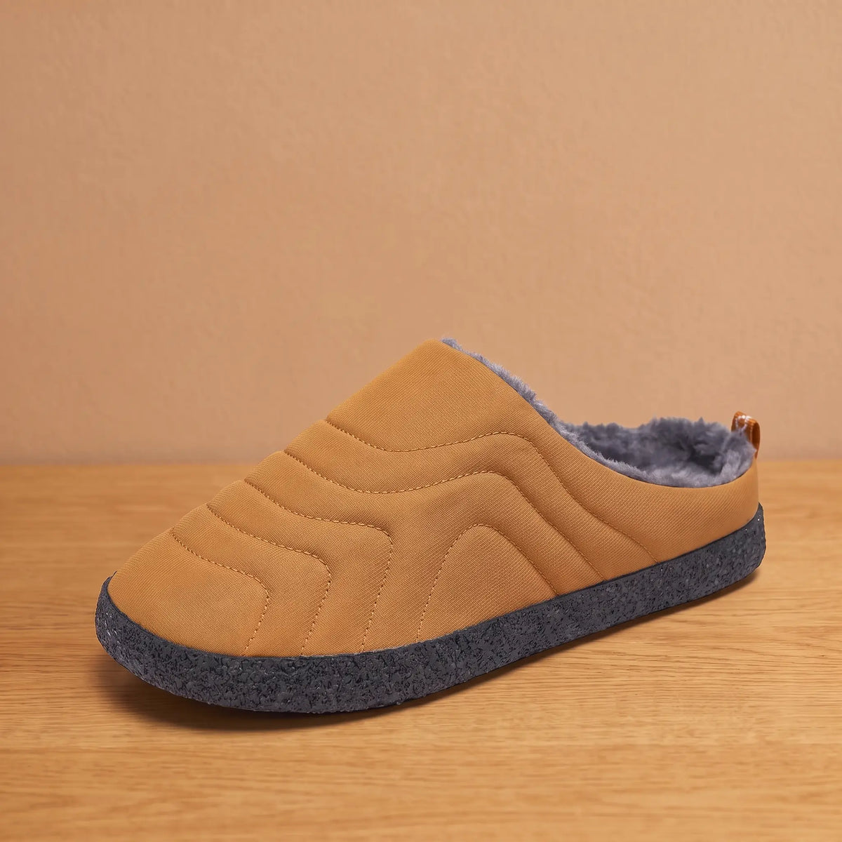 Men's khaki quilted backless slipper with plush faux-fur lining and speckled sole on an oak floor