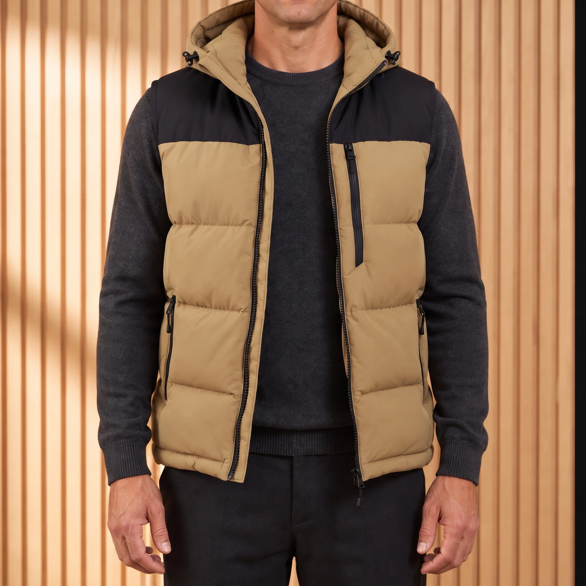 Men's khaki hooded puffer vest with black yoke, worn open over a charcoal knit