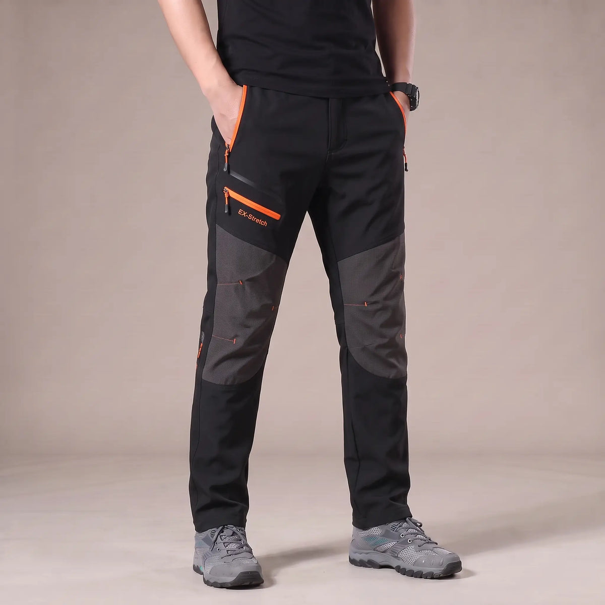Men's black stretch hiking trousers with dark grey knee panels and orange zip pockets, worn with hiking shoes