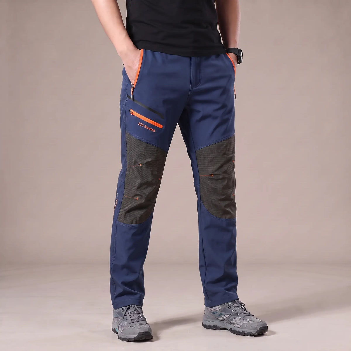 Men's blue stretch hiking trousers with dark grey knee panels and orange zip pockets, worn with hiking shoes