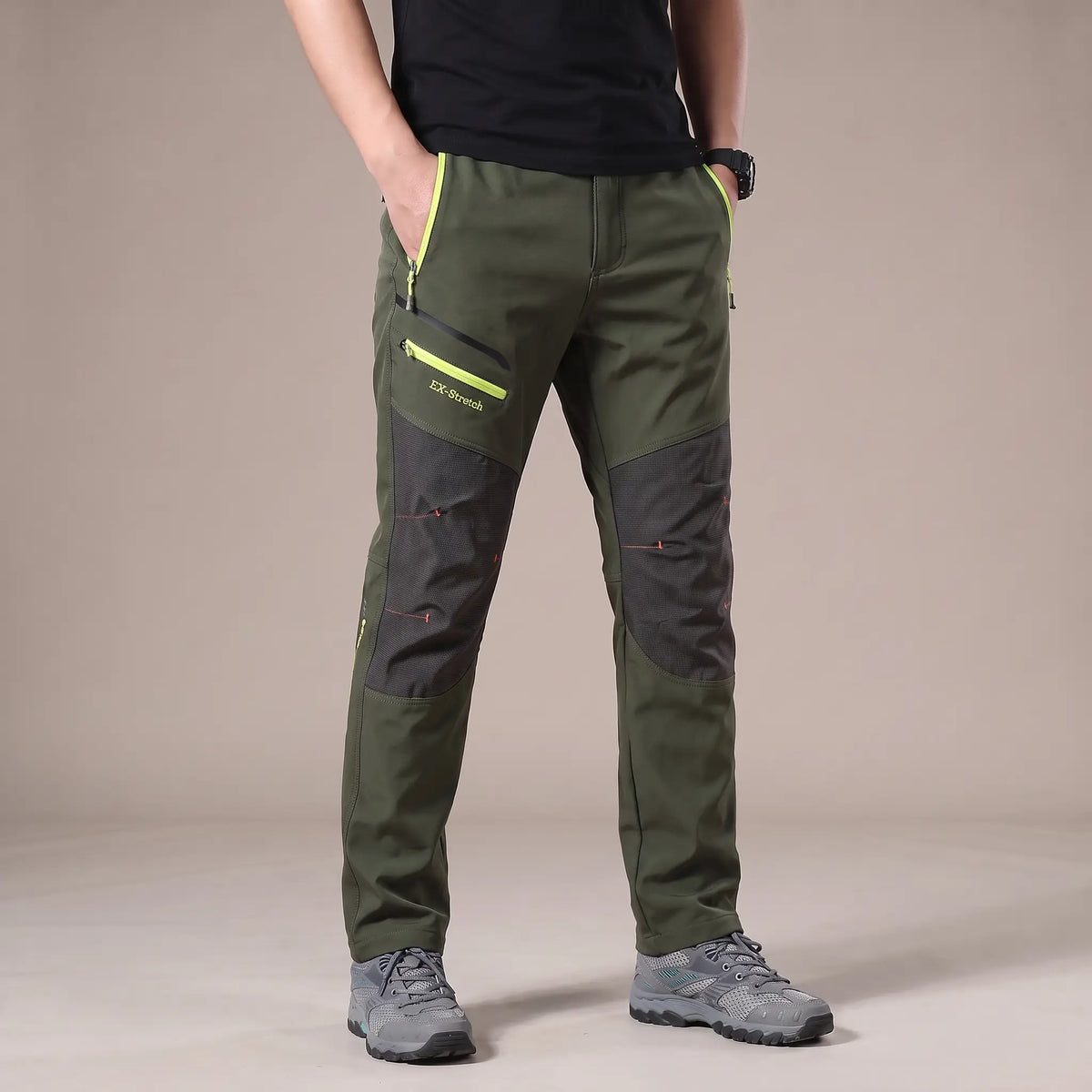 Men's green stretch hiking trousers with dark grey knee panels and lime zip pockets, worn with hiking shoes