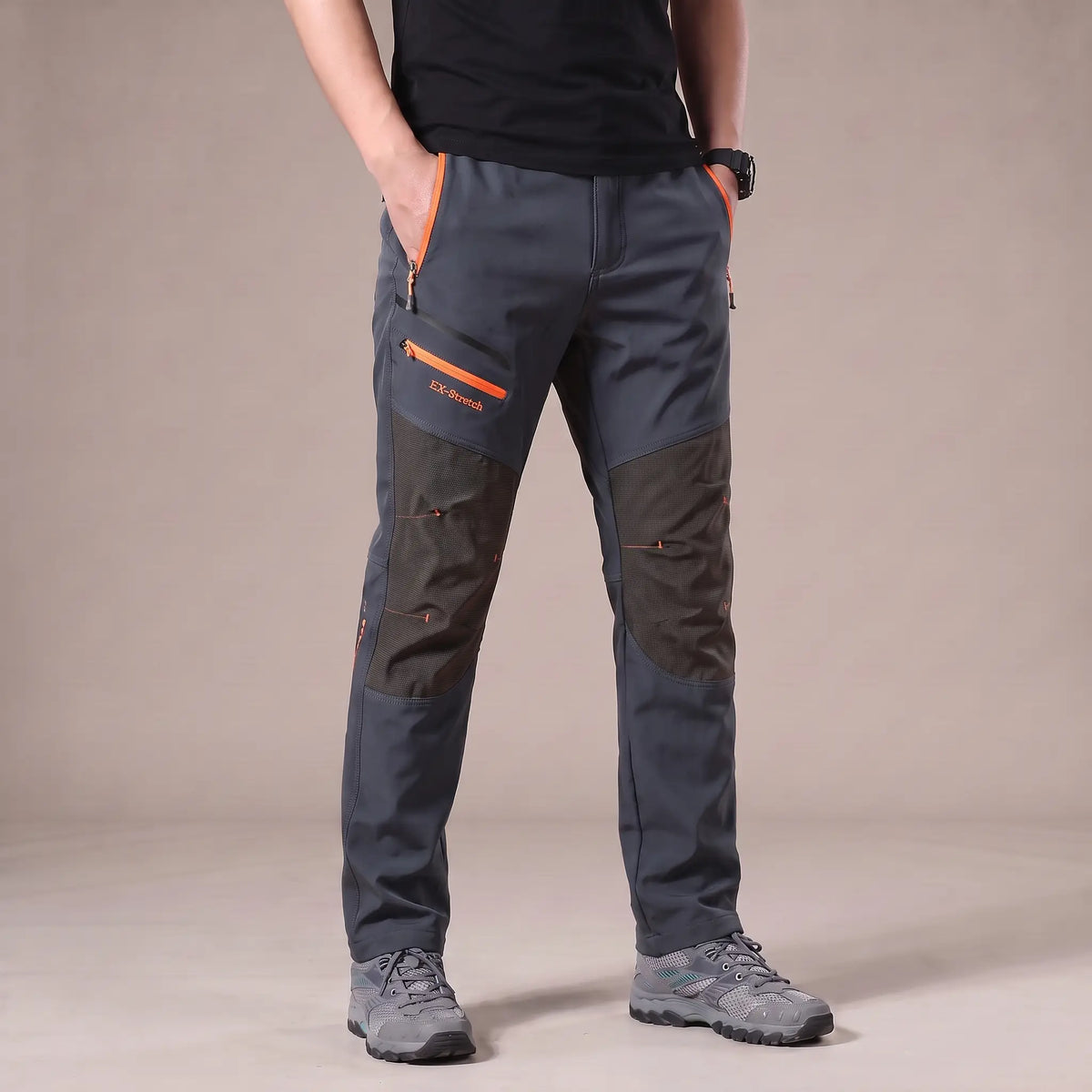 Men's grey stretch hiking trousers with dark grey knee panels and orange zip pockets, worn with hiking shoes