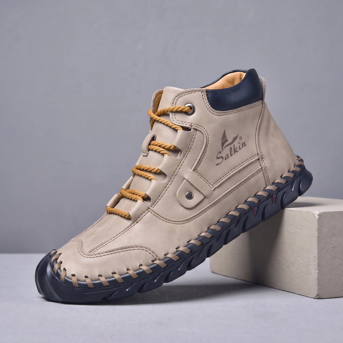 Beige hand-stitched men's lace-up ankle boot with navy sole, side view on a studio plinth