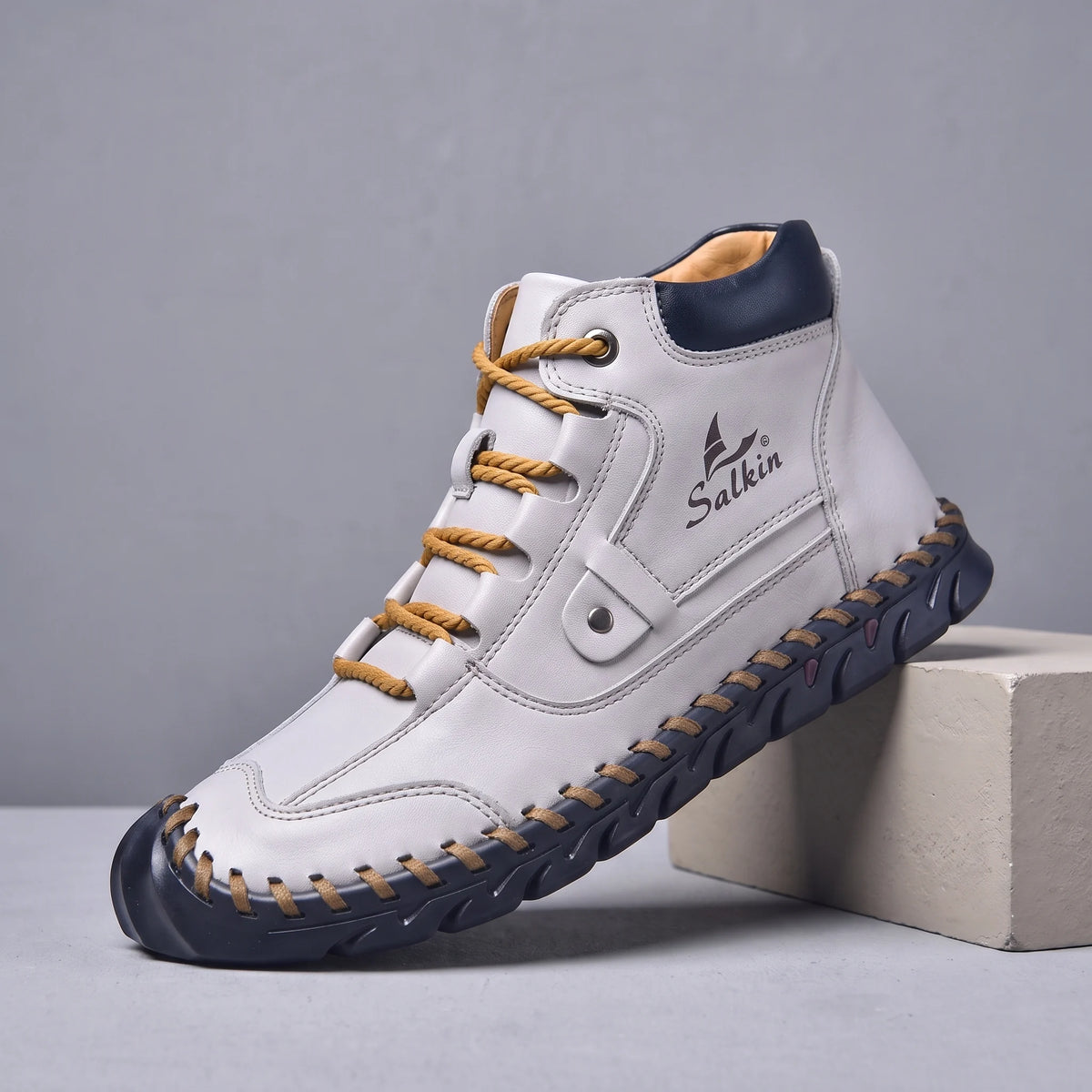 White hand-stitched men's lace-up ankle boot with navy sole, side view on a studio plinth