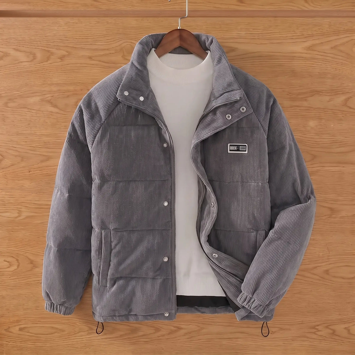 Grey padded corduroy puffer jacket on a wooden hanger against an oak plank wall