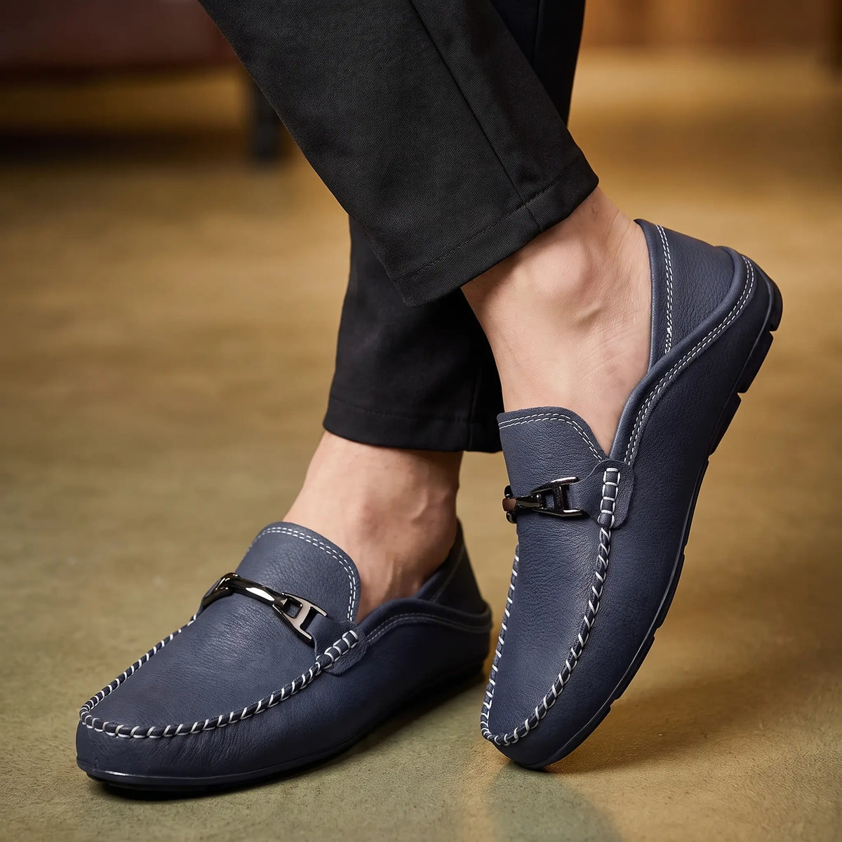 Man wearing blue pebbled leather driving loafers with dark trousers seated with ankles crossed