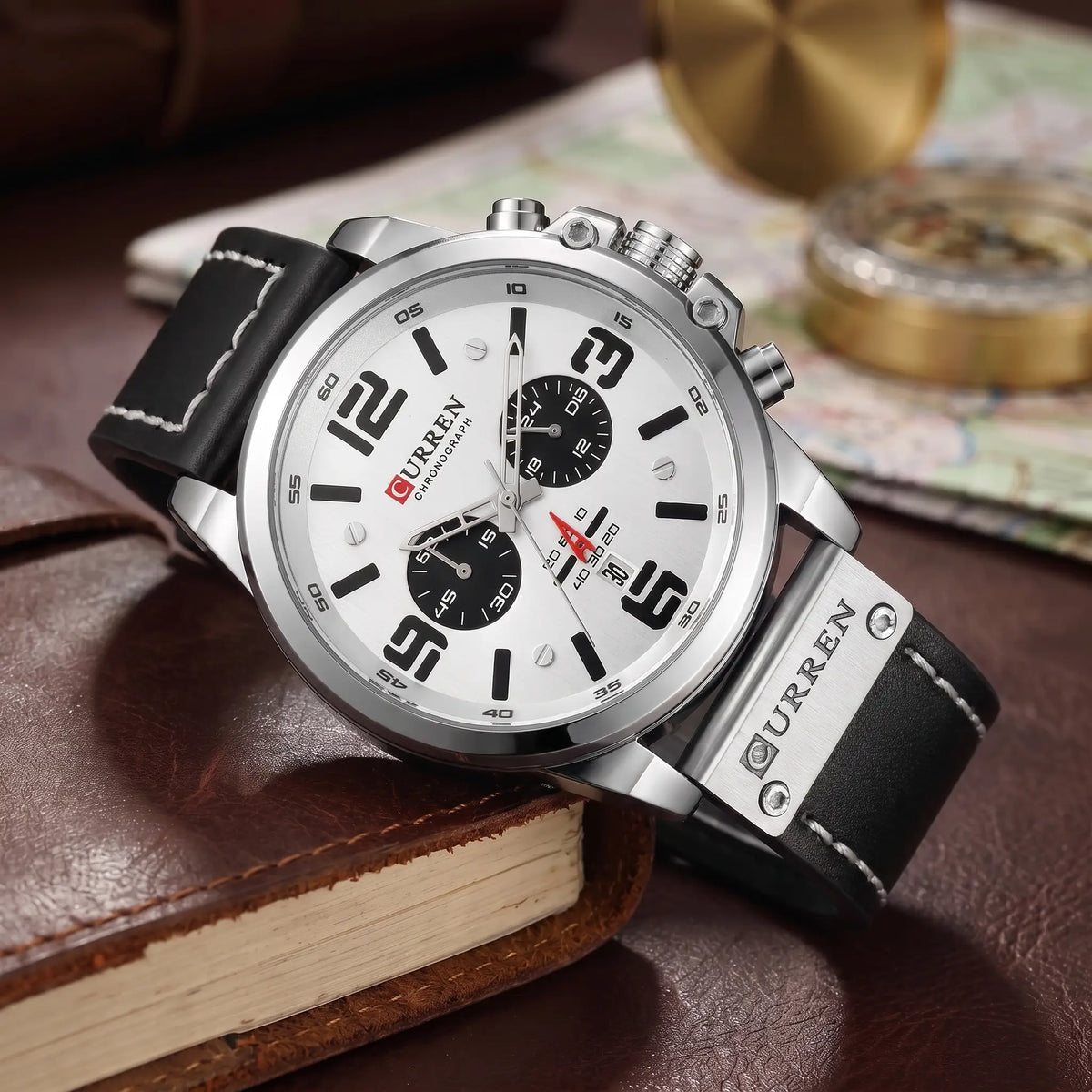 Silver chronograph watch with white dial and black leather strap draped over a leather journal