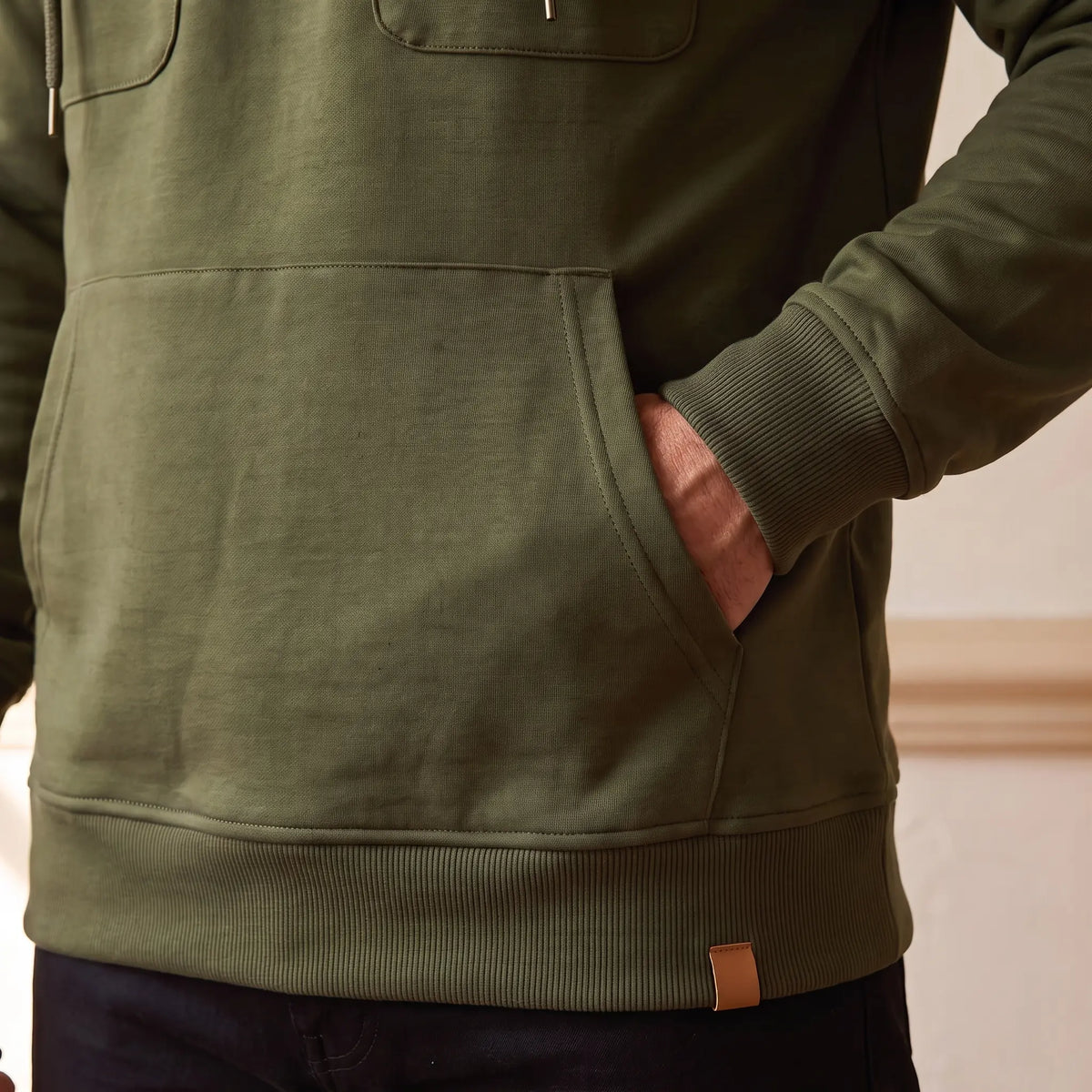 Close-up of the kangaroo pocket, ribbed hem and leather tab on the olive green hooded anorak