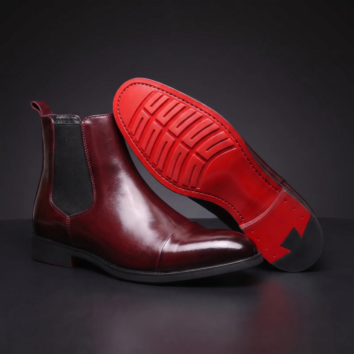 RED SOLE BOOTS burgundy leather Chelsea boot propped toe-up showing the detailed red lugged sole, dark charcoal scene