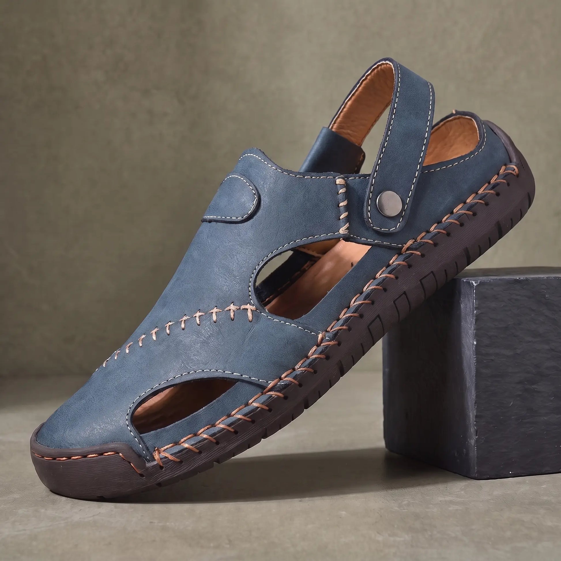Snap Strap Sandals in blue, a closed toe leather fisherman sandal with cut out side panels and a metal snap instep strap, on a charcoal slate block