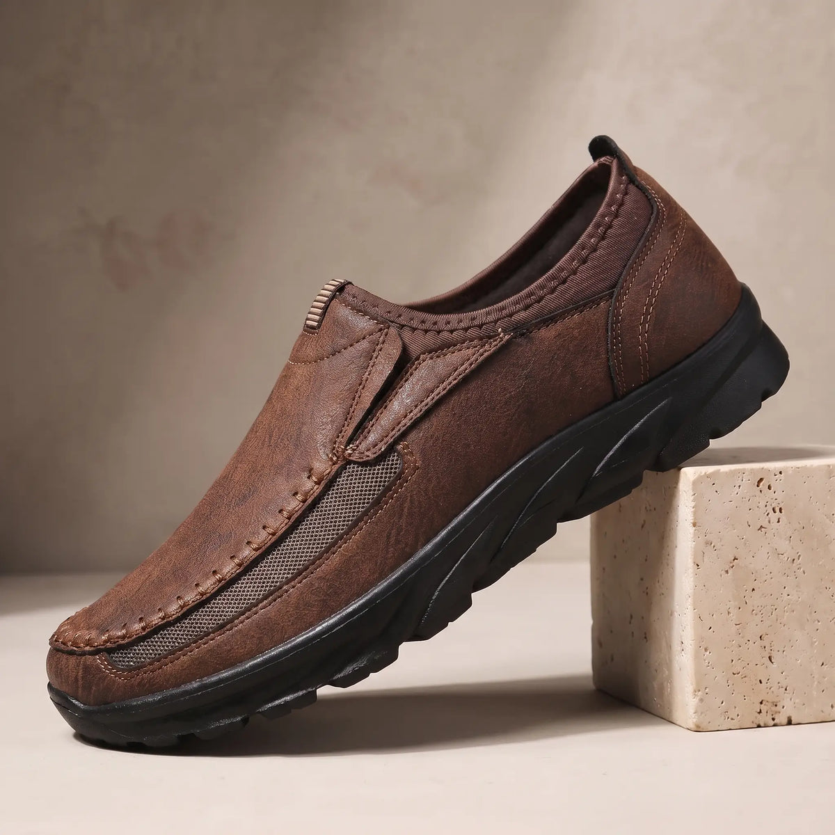 Stitched Apron Loafers in dark brown, a slip on moccasin loafer with elastic side panels and a chunky black bottom, on a travertine display block
