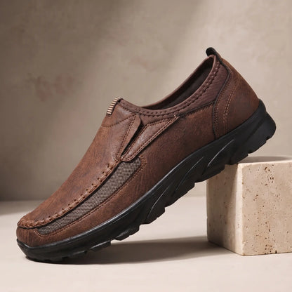 Stitched Apron Loafers in dark brown, a slip on moccasin loafer with elastic side panels and a chunky black bottom, on a travertine display block